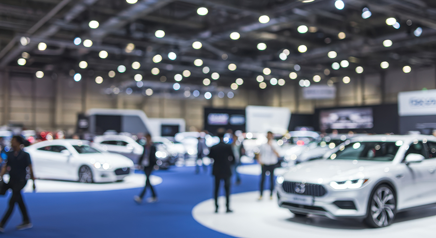 Blurry, large indoor exhibition hall filled with automotive vehicles and displays, featuring attendees in a commercial environment. The view is deliberately out-of-focus, conveying a sense of the bustling atmosphere of a motor vehicle show.